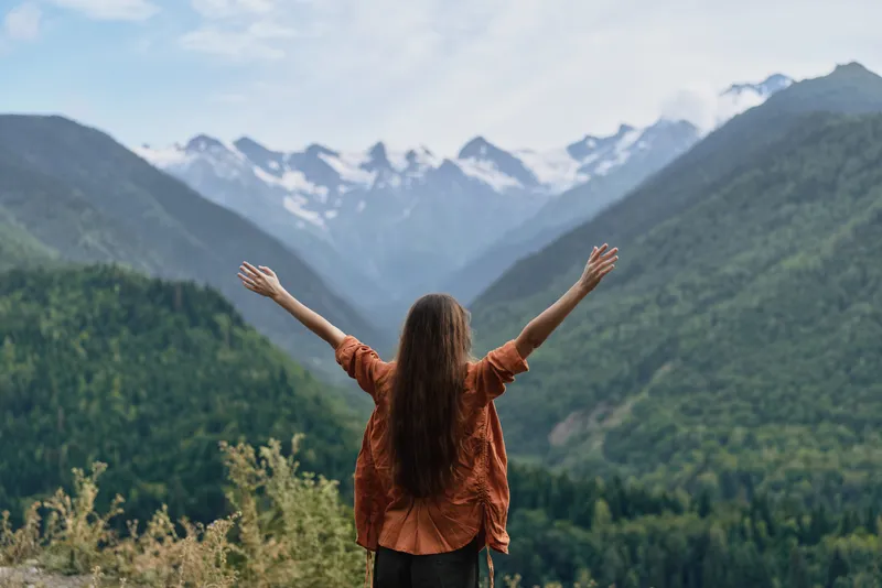 Woman with arms raised in a vast mountain landscape, embracing nature and freedom, gazing over green valleys toward snowy peaks under a clear alpine sky