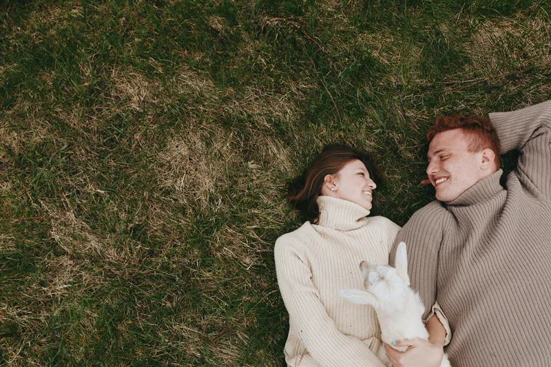 Couple outdoors, lifestyle scene with a goat kid, cuddling and smiling on grass, sunlit moment, cozy sweaters, relaxed pose, natural colors, breeze and greenery, intimate connection, playful and calm