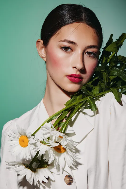Close-up of fashionable young woman with red lips holding white daisies in vibrant beauty and creative fashion photo concept on mint background.