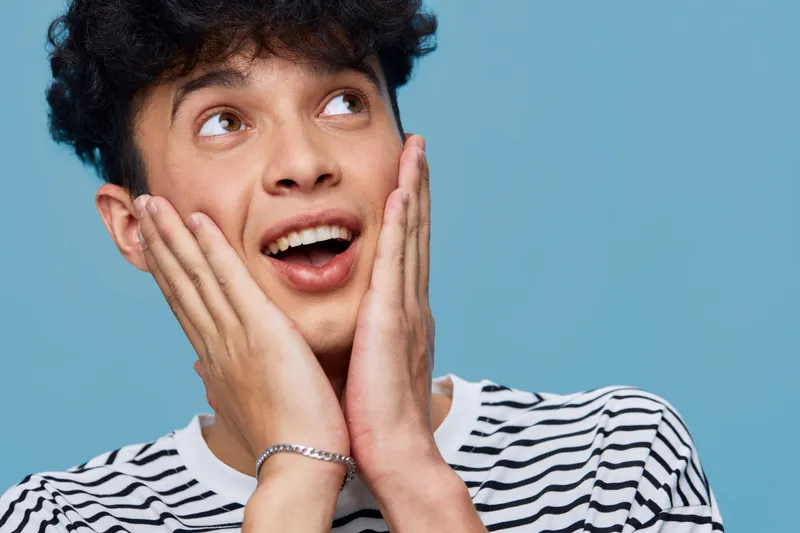 surprised man in striped shirt expressing amazement with hands on cheeks against blue background, capturing youthful energy and spontaneous emotion