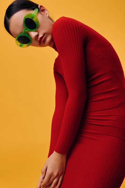 Woman in green sunglasses and red ribbed dress posing on yellow background for fashion, style, glamour and modern outfit in studio photo shoot with confident expression and trendy look.