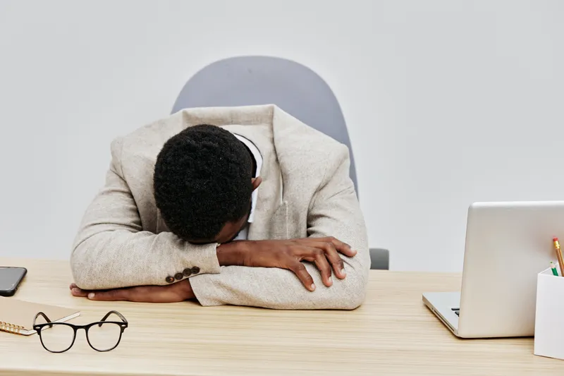 Frustrated young man with dark skin and short curly hair, wearing beige blazer, resting his head on desk while working on laptop, surrounded by glasses and notebooks, indoor office scene, serious mood