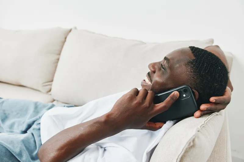 Young African man happily talking on the phone while relaxing on a couch, showcasing a casual lifestyle in a light and airy living room environment
