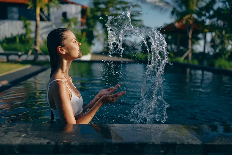 A woman sits in a pool with water spraying from her hands, eyes closed, savoring a moment of calm. Outdoor poolside scene conveying relaxation, leisure, and wellness in a sunny daytime setting.
