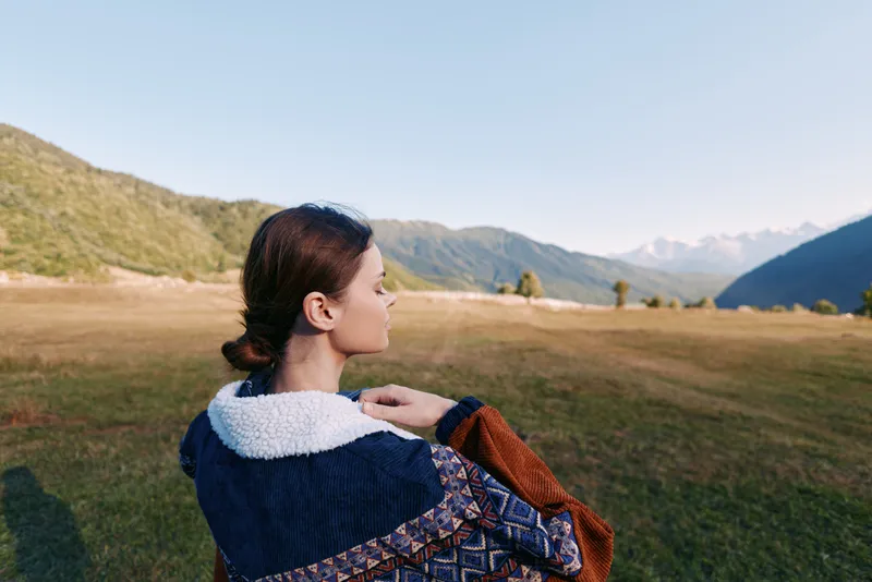 Woman in a warm jacket with fleece collar stands in a wide meadow overlooking distant mountains, profile view outdoors. Travel portrait capturing landscape, calm horizon and soft autumn light.