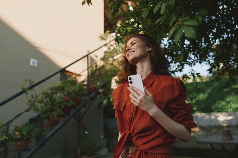 Cheerful woman outdoors wearing red, smiling with eyes closed, sunlit moment, relaxed posture, soft breeze, natural colors, lush greenery background, casual fashion, carefree mood, midday light