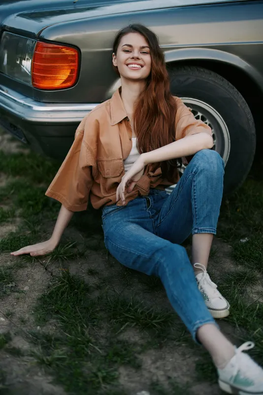 Young woman with long hair in casual outfit sitting on the grass next to a classic car, radiating joy and confidence in a sunny outdoor setting.
