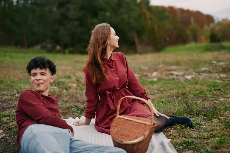Candid picnic scene of a young couple lounging on a blanket in a grassy meadow, showing genuine connection and authentic relaxed companionship with a wicker basket and soft natural light.