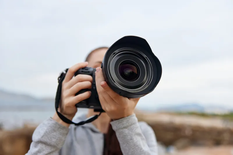 Young female photographer holding a camera on a cloudy day, showcasing passion for photography, with a blurred natural background emphasizing creativity and focus