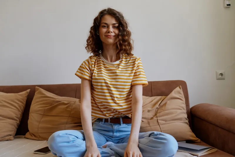 Smiling woman with curly hair sits crosslegged on a sofa in a cozy living room, reflecting a warm home lifestyle, comfort, and relaxed everyday moments.