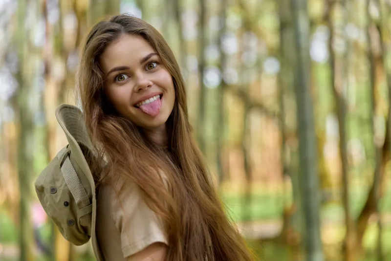 Woman portrait playful in forest showing tongue and smiling, longhair flowing, holding hat over shoulder, casual outdoor lifestyle, youth and natural light with carefree expression