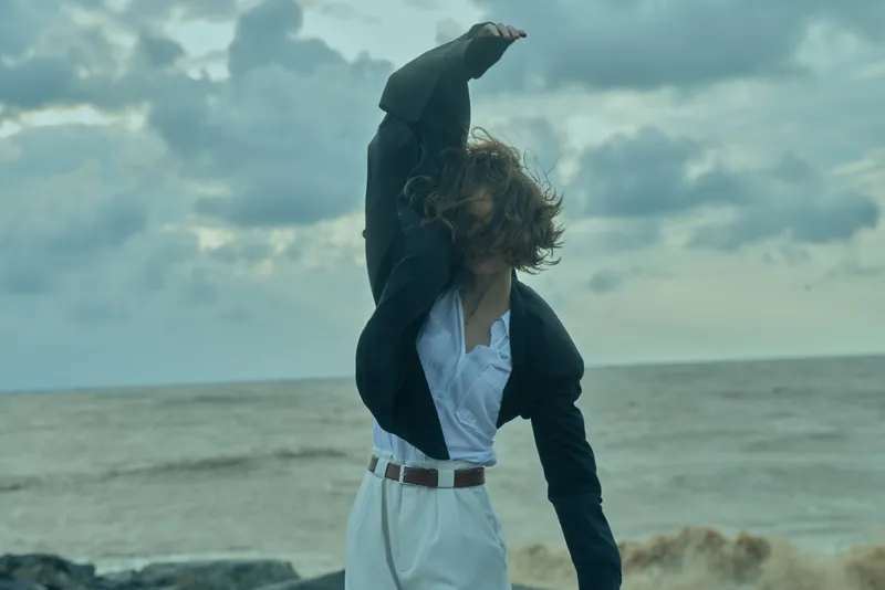 Woman in a smart blazer and white pants raises her arm against a windy seaside backdrop, expressing freedom, movement, and chic style by the ocean