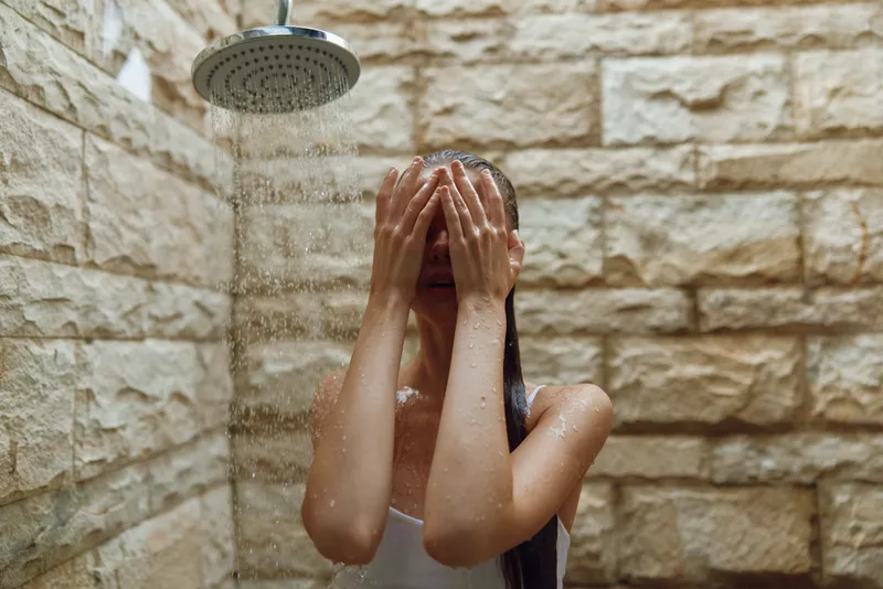 young woman washing face under rain shower with wet hair in modern stone bathroom, refreshing skincare routine, natural light and calm mood