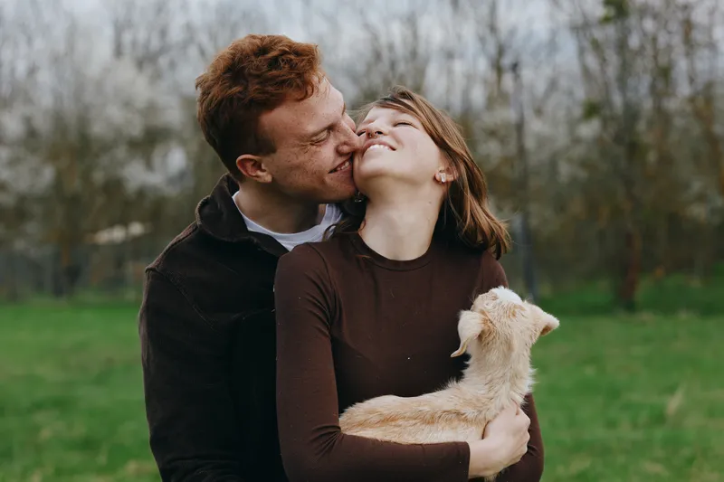Smiling couple embracing a playful goat kid in a green park, warm affection and natural light, lifestyle scene blending love and animal companionship, candid moment of joy and togetherness