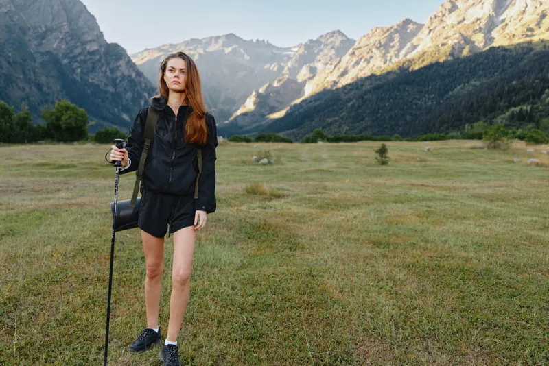 A young female hiker stands in a wide meadow with distant mountains, equipped with a trekking pole and backpack, conveying a calm, adventurous outdoor moment in natural scenery.