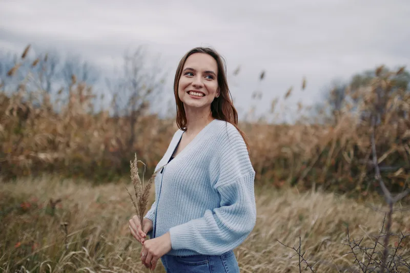 A smiling young woman in a cozy knit cardigan stands in tall grass, an authentic, genuine portrait capturing a candid outdoor moment with natural expression and relaxed posture.