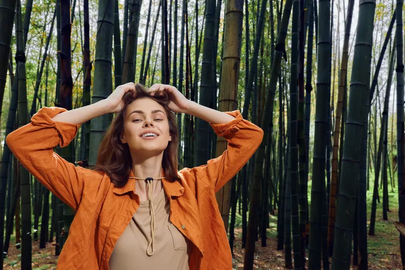 Woman bamboo forest smile portrait, young woman standing in a bamboo grove with hands in her hair, wearing an orange jacket, relaxed expression, natural outdoor beauty and morning light.