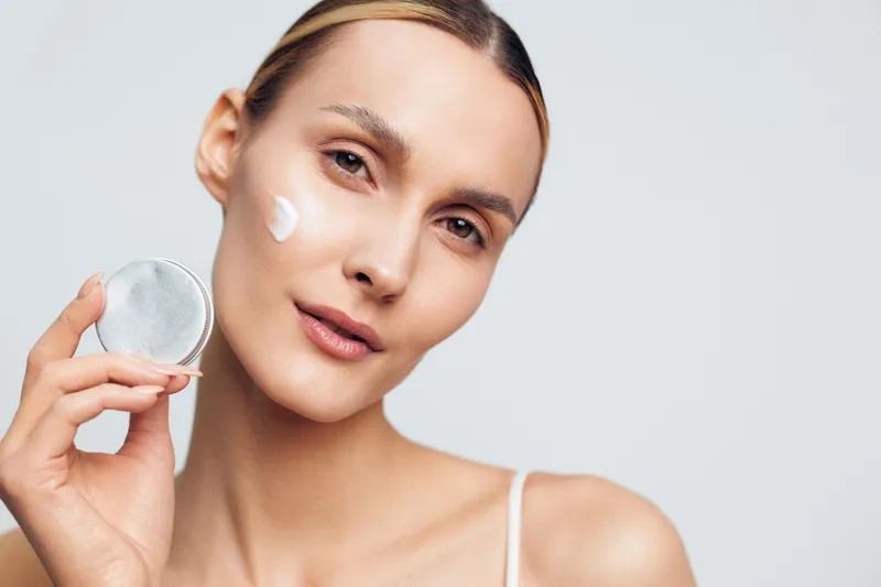 A young woman applies moisturizing cream on her face while holding an open jar. The close-up shows clear skin and natural beauty with a neutral background.