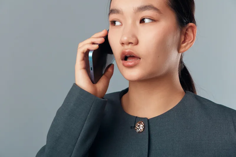 Young woman talking on smartphone with thoughtful expression wearing formal jacket for communication technology and business concept on plain background.