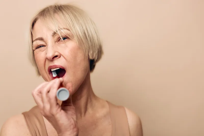 Mature woman with short blonde hair applies lipstick in a close-up shot on beige background. Concept of makeup routine, beauty care, and self-expression for older adults.