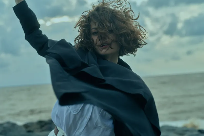 Wind swept outdoor scene with a carefree woman spinning by the sea, wearing a flowing jacket and white top, capturing motion and freedom