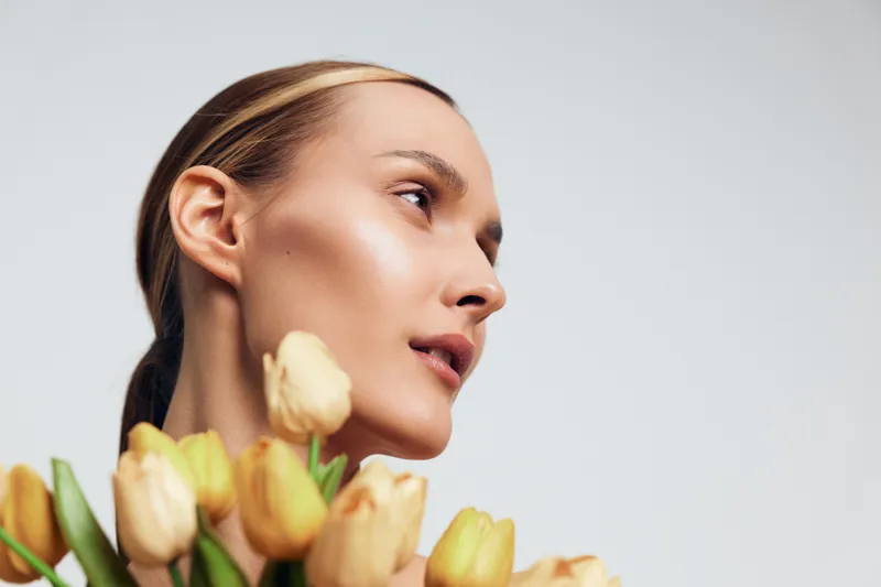A young woman with clear skin and pulled-back hair holds a bouquet of yellow tulips, looking thoughtfully to the side against a plain light background, symbolizing natural beauty and calm.