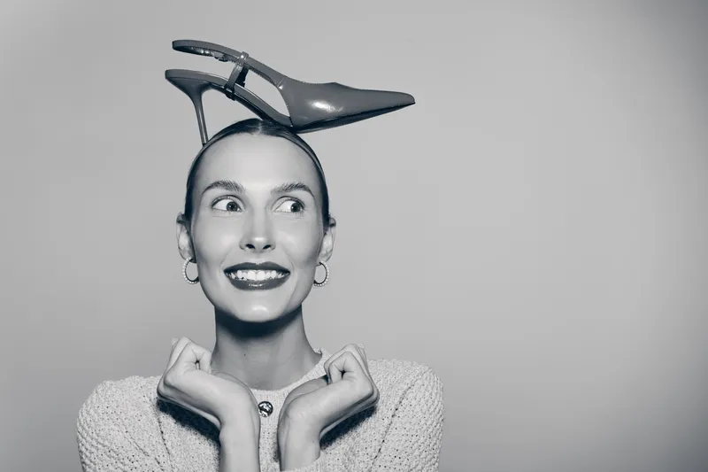 Smiling Caucasian woman with light skin balances high heel shoe on head in studio. Concept for fashion, fun, beauty.
