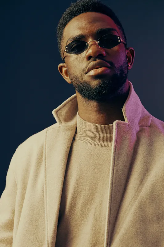 Stylish Black man wearing sunglasses and a beige coat, posing confidently against a dark background, highlighting modern fashion trends and contemporary style