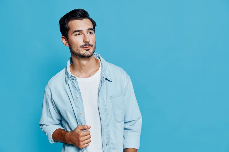 Young thoughtful man in light blue shirt gazing to the side with a calm expression on his face, standing against a bright turquoise background. Lifestyle concept