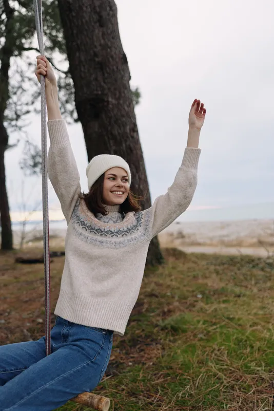 Woman on a swing in an open outdoor setting wearing a cozy sweater and beanie, smiling and carefree as she enjoys nature, trees and a breezy day near the coast