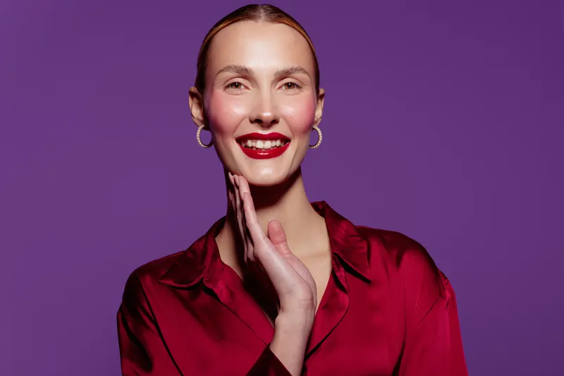 Smiling woman with red lipstick and earrings wears red satin blouse on purple background. Portrait of confident female model with hand near face. Concept of beauty, fashion, and elegance.