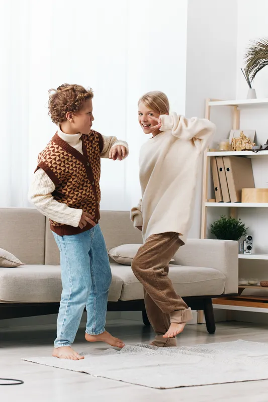 Two children dance playfully in a bright living room, smiling and carefree as they move together, wearing cozy knitwear and soft neutral tones during a sunny afternoon.