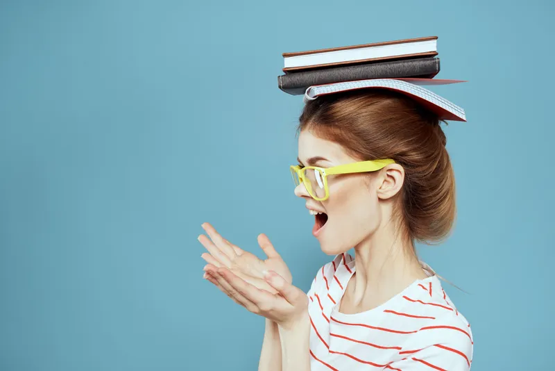 Студентка держит книги на голове и в жёлтых очках на синем фоне, обрезанный вид