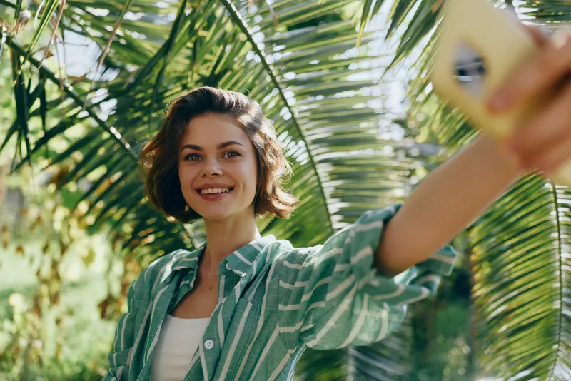 Young woman smiles while taking a selfie in a sunny tropical garden, green palm leaves framing a bright candid moment.