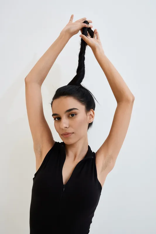 young woman in black sleeveless top tying long dark hair, natural look, minimalist background, casual style, indoor portrait with soft lighting