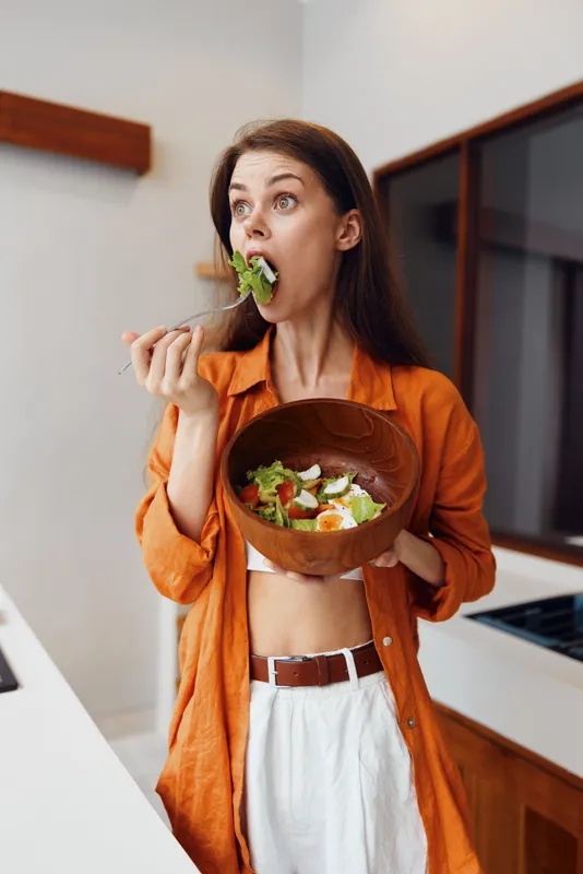 Молодая женщина в оранжевой блузке со страстью наслаждается вкусным салатом в деревянной миске на фоне светлой кухни