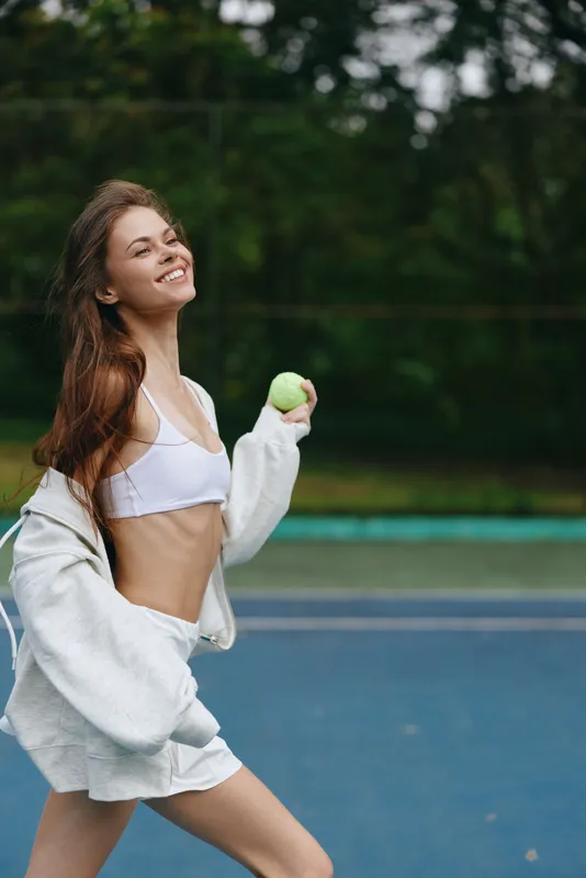 Young woman in sporty outfit smiling and holding a green apple on a tennis court, epitomizing healthy lifestyle and fitness concepts, showcasing joy and vitality