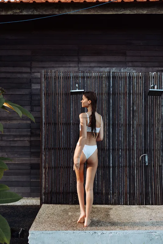 Slim woman in white bikini standing outdoors under outdoor shower, wet hair, wooden wall background, summer lifestyle, natural light, relaxed mood, vacation setting