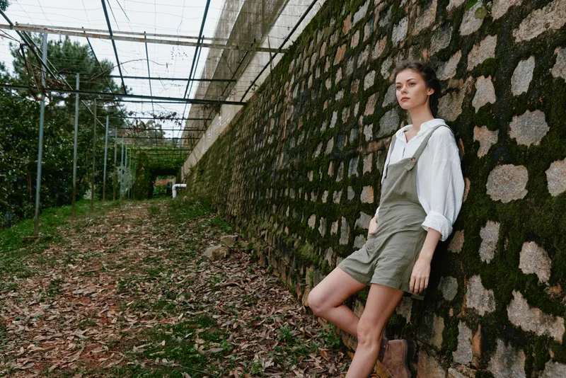 Young woman in casual green overalls and a white shirt, standing against a stone wall in a nature filled environment, evoking a sense of tranquility and connection with nature