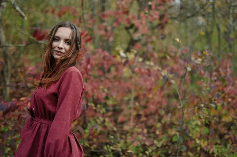 Young woman in a simple burgundy dress stands in autumn woodland, captured in an authentic candid portrait that highlights genuine expression, natural light and credible everyday beauty and presence.