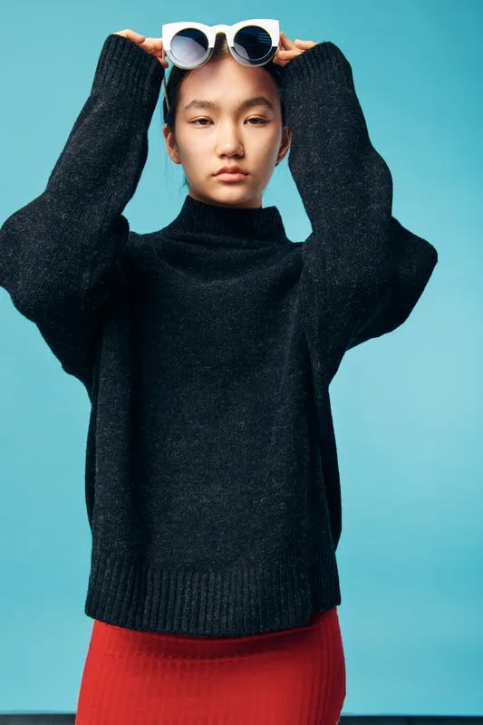 Young woman in black sweater and red skirt holding round white glasses on her head looking at camera with calm expression against blue background studio portrait.