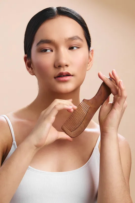 Asian woman gently brushes her hair with a wooden comb in a soft, neutral setting, exuding calm concentration and personal care in a minimal, intimate portrait.