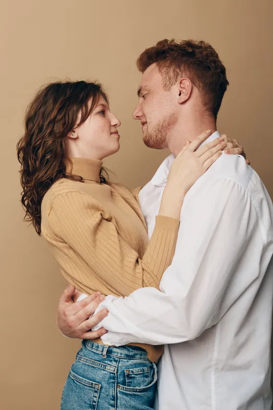 Young couple embraces closely looking into each other eyes with gentle expressions. Casual clothing and warm beige background create intimate romantic mood and connection between lovers.