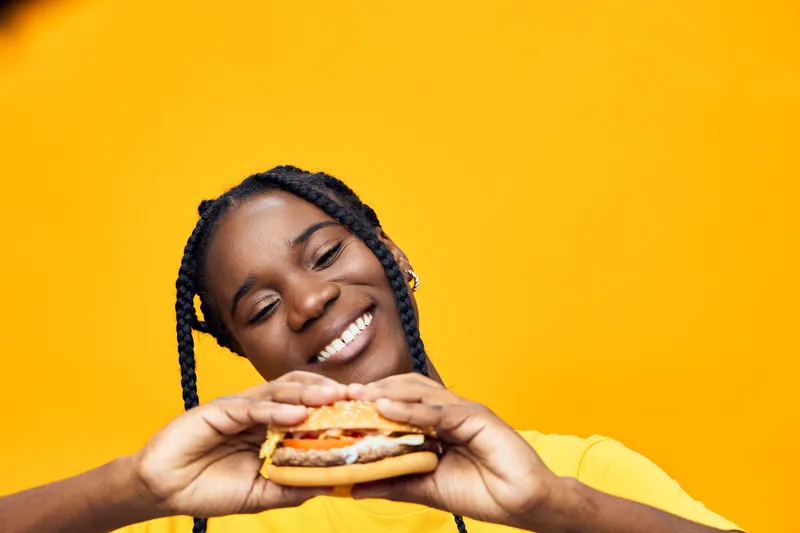 Joyful young woman of African descent holding a delicious burger against a vibrant yellow background, showcasing a fun and casual food atmosphere