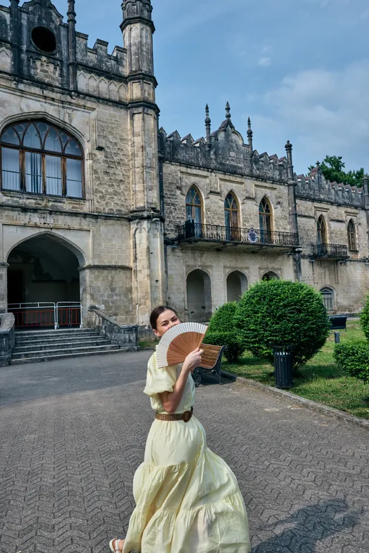 Elegant woman in a flowing pale yellow dress holds a fan as she poses outdoors in front of a medieval castle, creating a graceful timeless scene, bright mood and classic charm.