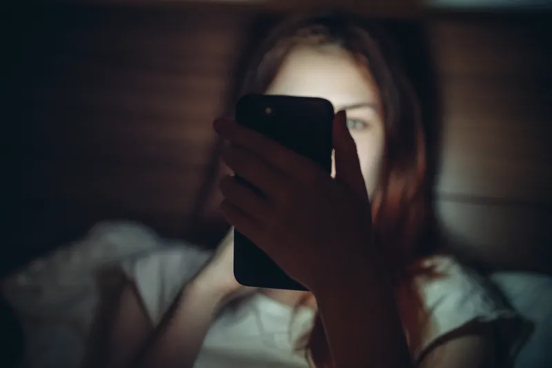 Девушка лежит в темноте и смотрит в светящийся смартфон