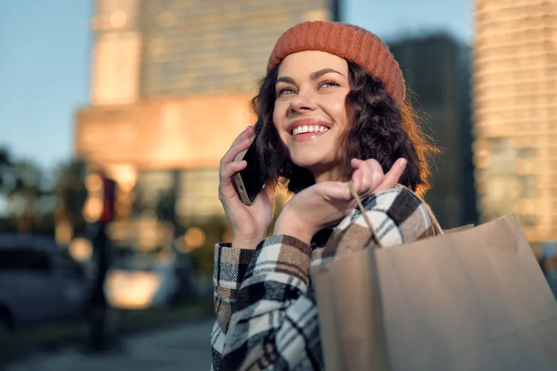 Woman shopping with phone and smile in city, candid lifestyle portrait showing authenticity and golden hour glow, mindful living moment of emotional storytelling and joyful connection.