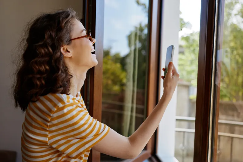 Woman at home by a window, smiling and holding a smartphone, casual striped t shirt, warm natural light, relaxed cheerful mood, everyday lifestyle indoors