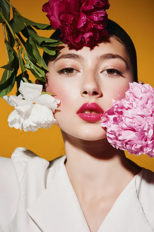 Close-up portrait of a young woman with vibrant glossy lips posing with bright peony flowers around her head. Expressive beauty concept with creative fashion makeup and colors.