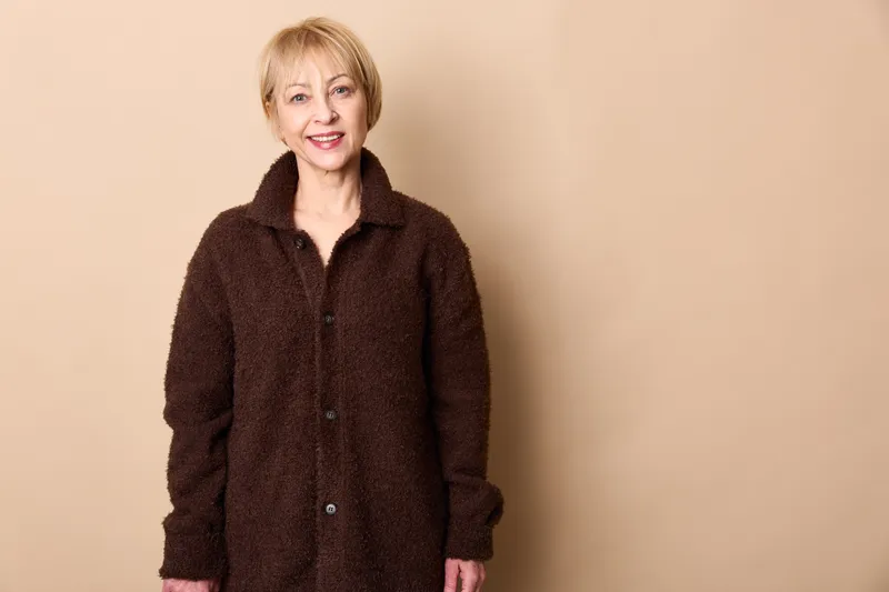 Woman portrait, smile, studio coat styling, blonde mature hair, beige background, headshot, friendly expression, warm soft light, relaxed indoor posture